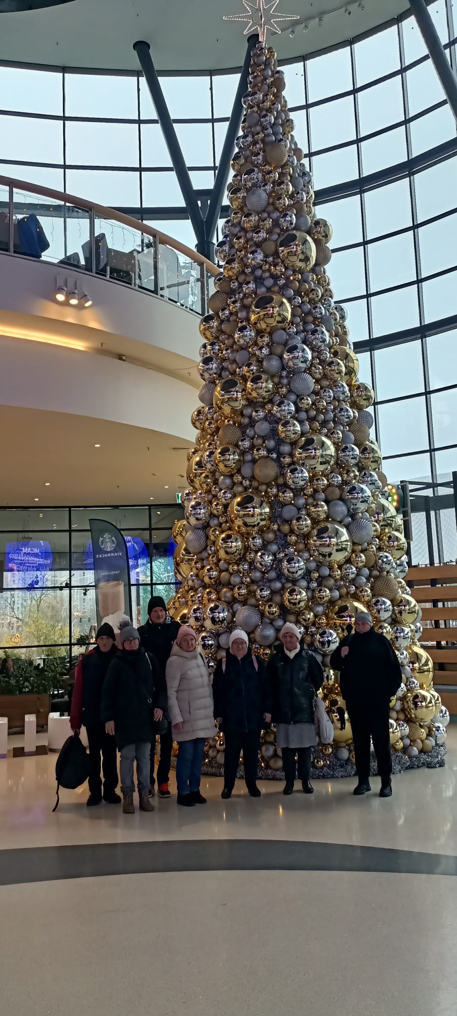 Uczestnicy ŚDS stoją do zdjęcia grupowego wewnątrz galerii Posnania. Za nimi stoi ogromna choinka wypełniona po brzegi złotymi i srebrnymi bombkami wszystkich rozmiarów