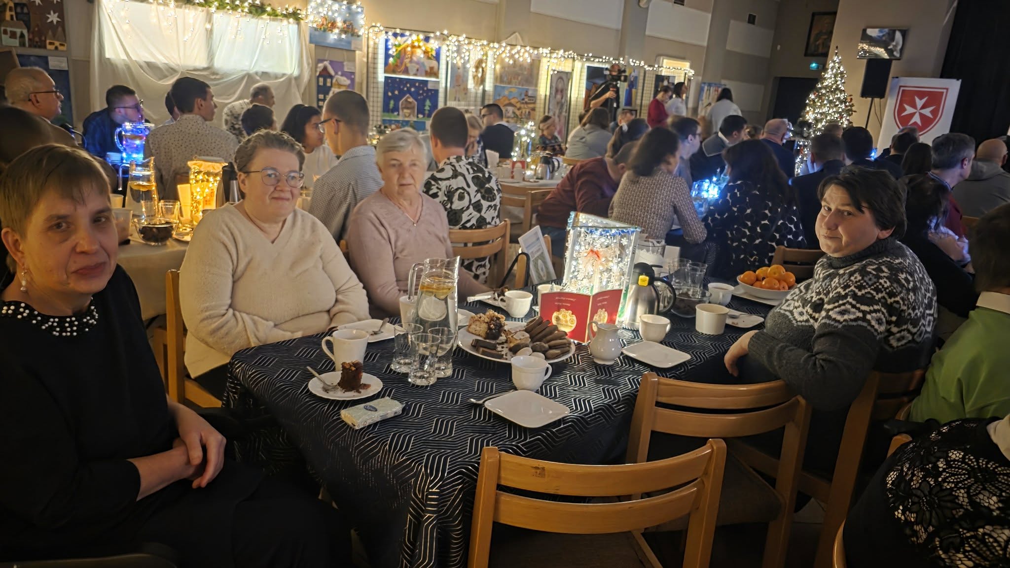 Uczestniczki ŚDS siedzą przy stole podczas spotkania. W tle widać sale udekorowaną światęcznie światełkami oraz choinkę