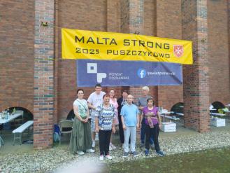 Grupowe zdjęcie uczestników Środowiskowego Domu Samopomocy przed budynkiem z czerwonej cegły. Na budynku zawieszone są dwa bannery: żółty z czarnym tekstem 'Malta Strong 2025 Puszczykowo' i herbem miasta oraz niebieski z białym tekstem 'Powiat poznański'