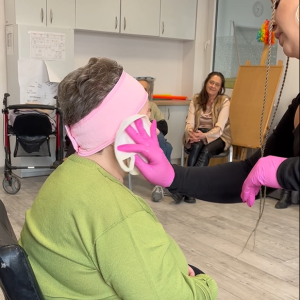 Uczestniczka ŚDS siedzi, a kosmetolożka w różowych nitrylowych rękawiczkach za pomocą akcesoriów masuje jej twarz