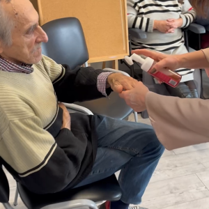 Uczestnik ŚDS siedzi z wyciągniętą dłonią, a kosmetolożka nakłada na nią preparat za pomocą specjalnego dozownika kosmetyku