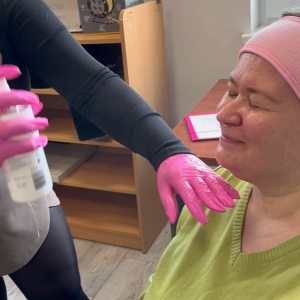 Uczestniczka ŚDS siedzi i mruży oczy podczas gdy kosmetolożka w różowych nitrylowych rękawiczkach spryskuje jej twarz za pomocą kosmetyku