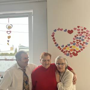 Trzech uczestników ŚDS pozuje do zdjęcia. Za nimi na ścianie i oknie wiszą papierowe ozdoby w kształcie serc.