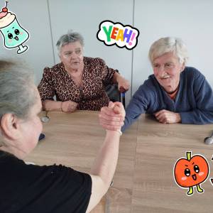 Mężczyzna i kobieta trzymają się dłońmi przy stole w pozycji jak gdyby mieli się siłować. Obydwoje są uśmiechnięci.