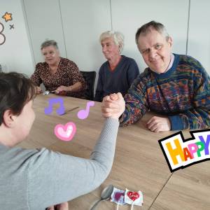 Mężczyzna i kobieta trzymają się dłońmi przy stole w pozycji jak gdyby mieli się siłować. Obydwoje są roześmiani.