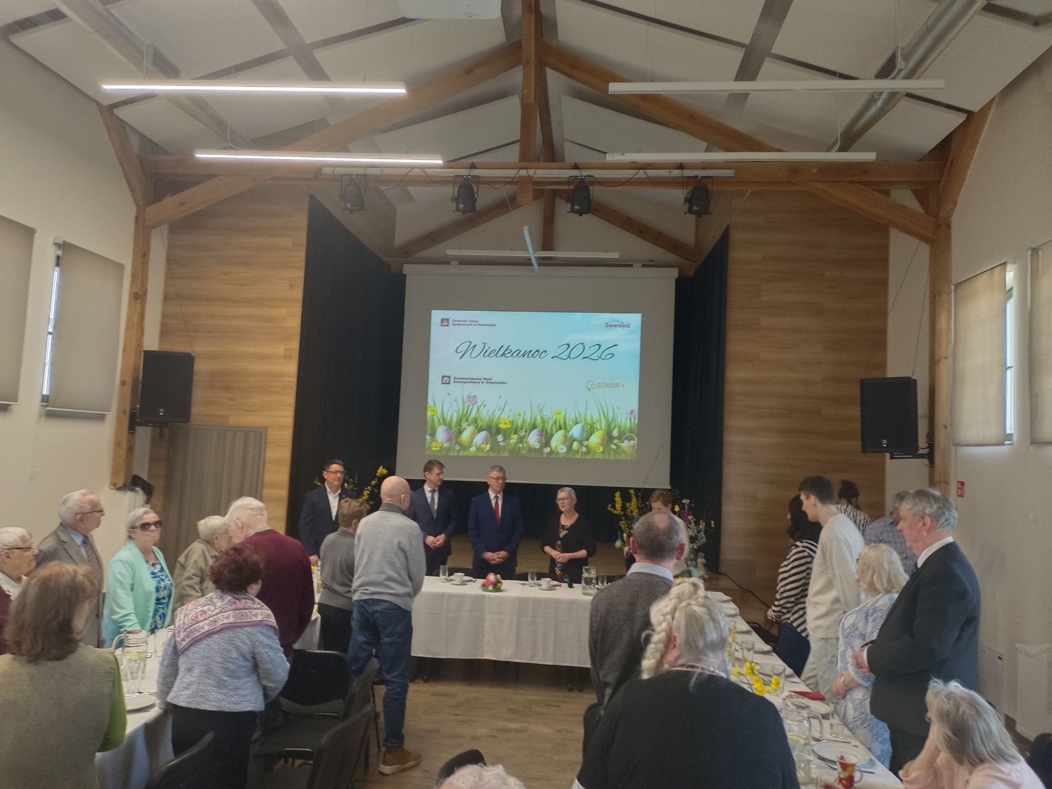 Zebranie osób w świetlicy. Na ścianie, za pomocą projektora, wyświetlana jest grafika Wielkanoc 2026 z logotypami organizujących spotkanie placówek. Za stołami z zastawą są uczestnicy ŚDS, Dzienniego Domu Pomocy Senior-Wigor oraz wybrane osoby z Gminy Swarzędz. 