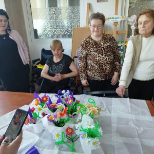 Uczestniczki ŚDS wraz z instruktorką podczas zajęć zebrały się przy stole. Na środku stołu stoją stworzone przez nich kolorowe bukiety stworzone z drucików florystycznych
