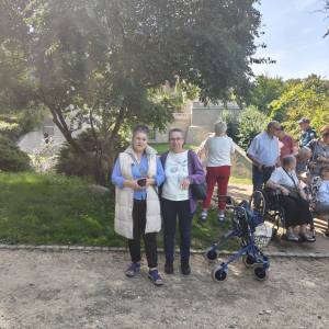 Dwie uczestniczki wycieczki do Gołuchowa stoją na parkowej ścieżce pod koroną rozłożystego drzewa, pozując do pamiątkowego zdjęcia. Jedna z kobiet ma na sobie jasną kamizelkę bez rękawów, a druga fioletowy kardigan i okulary. W tle po prawej stronie widać pozostałych członków grupy – seniorów spacerujących alejkami oraz osoby korzystające z wózka inwalidzkiego i chodzika rehabilitacyjnego. Wspólny wyjazd integracyjny został zorganizowany przez Dzienny Dom „Senior-Wigor” w Swarzędzu oraz Środowiskowy Dom Samopomocy.