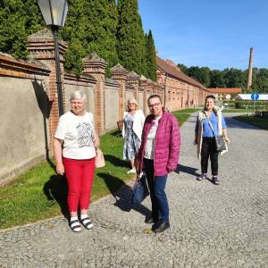 Uczestniczki wycieczki do Gołuchowa spacerują brukowaną alejką wzdłuż zabytkowego muru z czerwonej cegły w słoneczny dzień. Kobiety uśmiechają się do aparatu podczas wspólnego zwiedzania okolic zamku. W tle widoczne są historyczne zabudowania folwarczne, wysoki komin oraz zadbana parkowa zieleń pod bezchmurnym niebem. Wyjazd został zorganizowany wspólnie przez Dzienny Dom „Senior-Wigor” w Swarzędzu oraz Środowiskowy Dom Samopomocy.