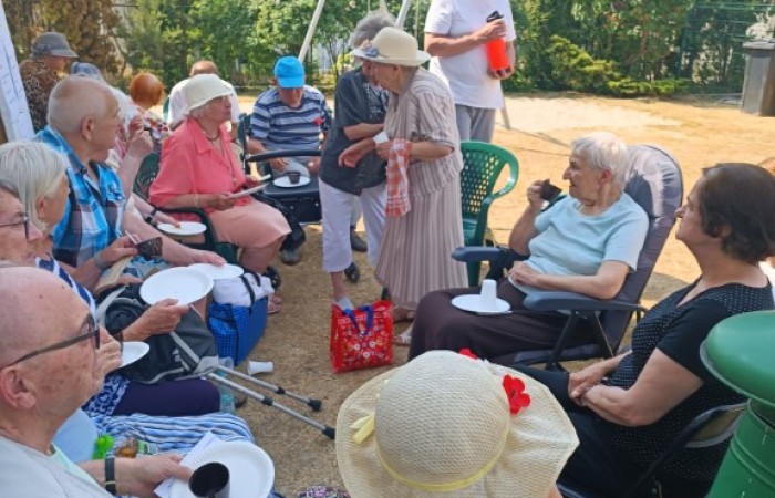 Grupa seniorów siedzi w kręgu na plastikowych krzesłach na podwórku, spożywając posiłki i rozmawiając. Mężczyzna w białej koszulce nalewa napój z termosu.