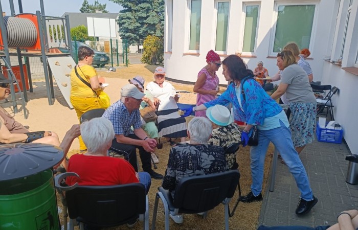 Grupa seniorów siedzi na krzesłach na zewnątrz, przy budynku z białymi ścianami i dużymi oknami. Część osób spożywa posiłki, inne rozmawiają. Wolontariusze i pracownicy w kolorowych ubraniach pomagają w obsłudze wydarzenia. W tle widać plac zabaw i ogrodzenie.