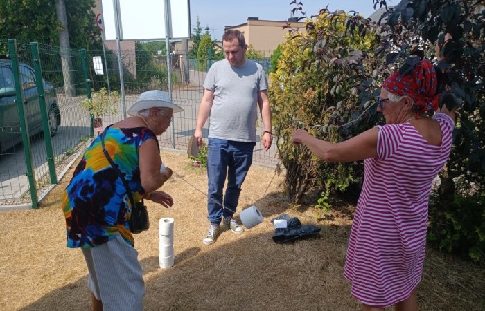 Dwie seniorki biorą udział w zabawie z rozwijaniem rolki papieru toaletowego. Mężczyzna w szarej koszulce nadzoruje grę, stojąc obok nich. Tło stanowią trawnik i ogrodzenie.