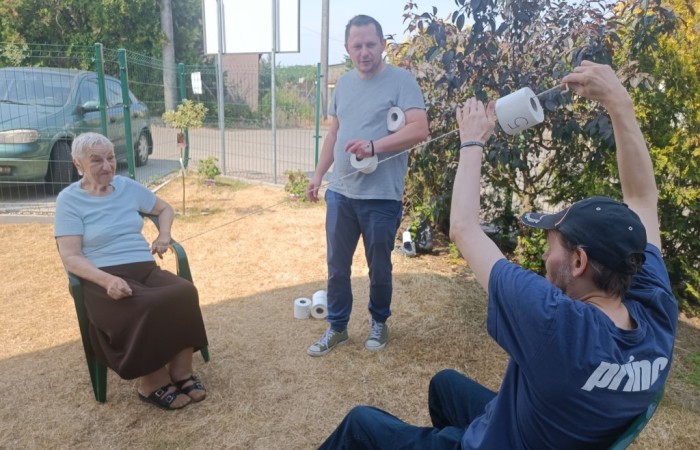 Starsza kobieta siedzi na plastikowym krześle, podczas gdy dwóch mężczyzn trzyma rolki papieru toaletowego na drewnianym kiju. Wydają się brać udział w grze na świeżym powietrzu.