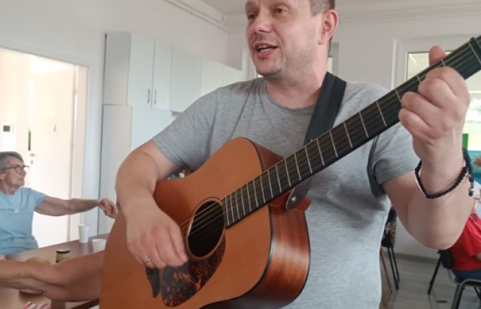 Mężczyzna w szarej koszulce i niebieskich spodniach gra na gitarze akustycznej w pomieszczeniu. Ma na twarzy wyraz zaangażowania, jakby śpiewał lub rozmawiał. W tle widać seniorów siedzących przy stole, z których jedna osoba wyciąga rękę w geście rozmowy lub kontaktu z inną osobą. Pomieszczenie jest jasne, z białymi szafkami i dużymi oknami.