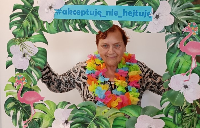 Starsza kobieta uśmiecha się i pozuje do zdjęcia, trzymając dużą ramkę udekorowaną zielonymi liśćmi, białymi kwiatami i różowymi flamingami. Na ramce widnieje napis #akceptuję_nie_hejtuję. Kobieta ma na szyi wielokolorowy naszyjnik z kwiatów.