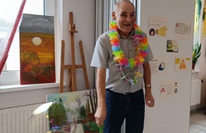 Starszy mężczyzna w kolorowym hawajskim naszyjniku stoi w jasnym pomieszczeniu, puszczając duże bańki mydlane. W tle sztalugi z obrazami oraz okno wpuszczające naturalne światło.