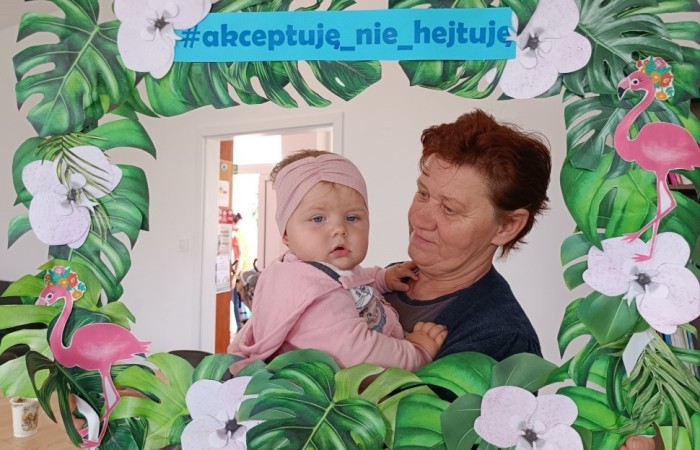 Starsza kobieta trzyma na rękach małe dziecko i obie pozują w dekoracyjnej ramce z tropikalnym motywem. Na górze ramki widnieje napis: #akceptuję_nie_hejtuję.