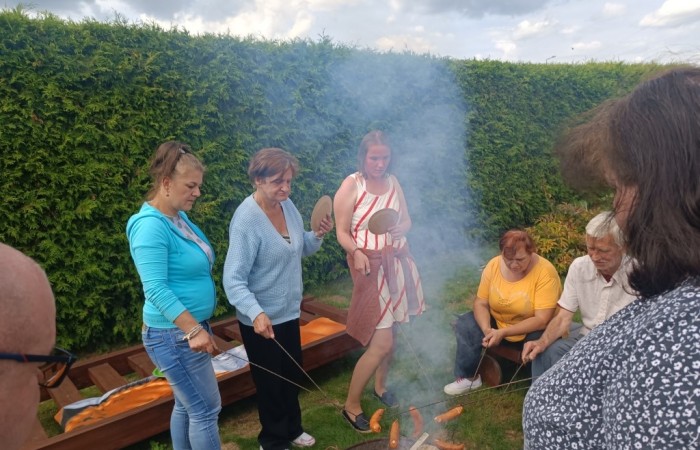 Kobiety pieką nabite na patyki kiełbaski nad ogniskiem, w tle żywopłot. Mają przygotowane papierowe talerzyki