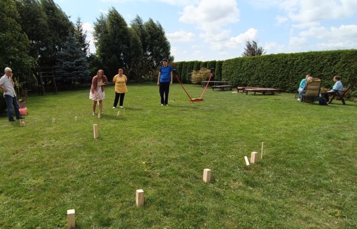 Grupa osób gra w grę na świeżym powietrzu, w ziemi powbijane są drewniane kołki. W tle na drewnianych ławkach, fotelach i krzesłach siedzą inne osoby.