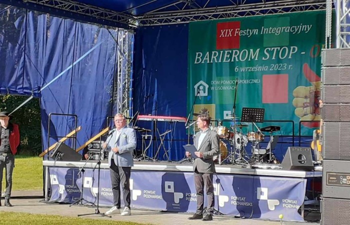 Zdjęcie przedstawia scenę plenerową podczas festynu integracyjnego „Barierom Stop” 6 września 2023 roku. Trzech mężczyzn stoi na scenie, jeden z nich przemawia do mikrofonu. W tle niebieskie zasłony sceniczne, baner festiwalu i sprzęt muzyczny, w tym perkusja i keyboard. Na pierwszym planie głośniki nagłaśniające