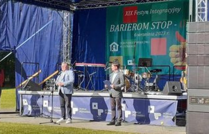 Zdjęcie przedstawia scenę plenerową podczas festynu integracyjnego „Barierom Stop” 6 września 2023 roku. Trzech mężczyzn stoi na scenie, jeden z nich przemawia do mikrofonu. W tle niebieskie zasłony sceniczne, baner festiwalu i sprzęt muzyczny, w tym perkusja i keyboard. Na pierwszym planie głośniki nagłaśniające.