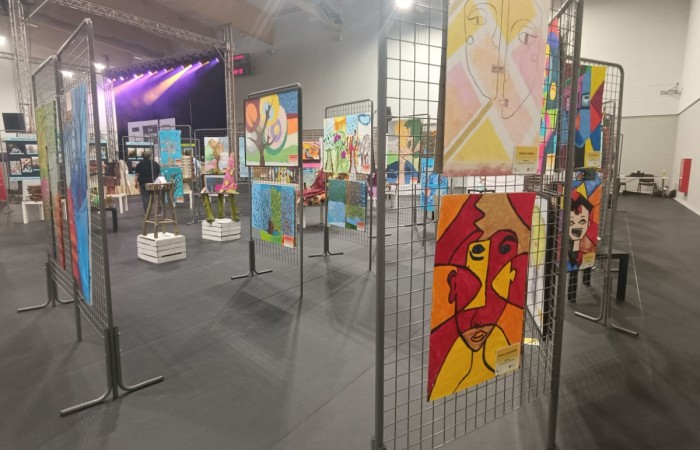 duża hala z ciemną podłogą, wypełniona metalowymi stojakami z siatki, na których wiszą kolorowe obrazy o abstrakcyjnych i geometrycznych kształtach. W tle scena z oświetleniem w odcieniach fioletu.