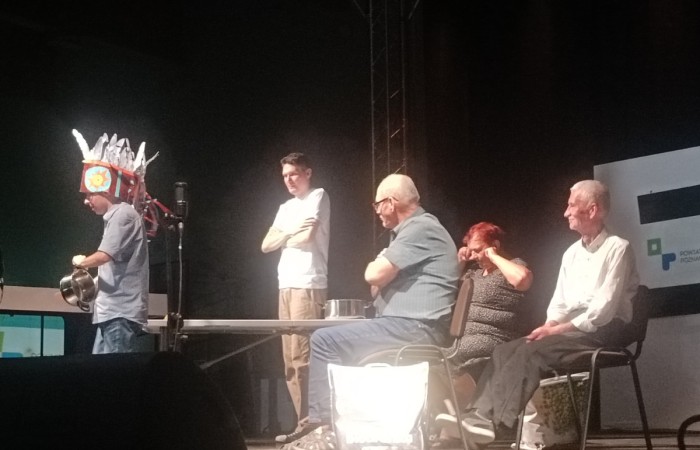 kilku starszych ludzi siedzi przy stole na scenie, patrząc na mężczyznę, który nosi nietypowy hełm z papierowych elementów i trzyma metalowe garnki. Jedna z osób na widowni wydaje się śmiać i ocierać oczy.