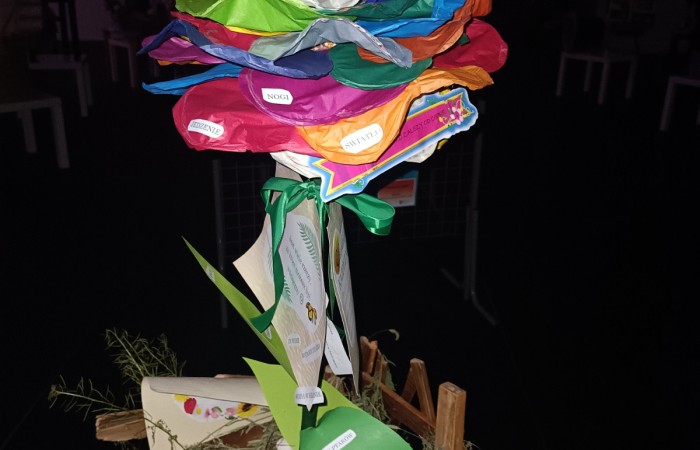 Kolorowa, ręcznie wykonana rzeźba przypominająca kwiat, wykonana z bibuły, papieru i innych materiałów. Płatki są w różnych kolorach – czerwonym, niebieskim, zielonym, pomarańczowym i fioletowym – i zawierają przyklejone napisy, takie jak „TALENT”, „NOGI” i „MYŚLENIE”. Łodyga składa się z owiniętego papieru, a przy podstawie znajdują się papierowe liście i karteczki z tekstami. Konstrukcja osadzona jest na drewnianej podstawce z elementami dekoracyjnymi, takimi jak siano i miniaturowe drewniane płotki. Tło jest ciemne, a rzeźba oświetlona jest błyskiem aparatu.