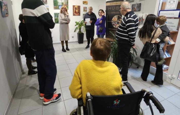 grupa ludzi zgromadzona w przestrzeni wystawy, w tym osoba na wózku inwalidzkim i rodzina z dzieckiem.