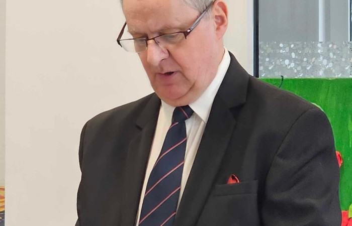 starszy mężczyzna w ciemnym garniturze i krawacie trzyma zieloną teczkę i odczytuje tekst. W tle biała ściana i zielona dekoracja. 