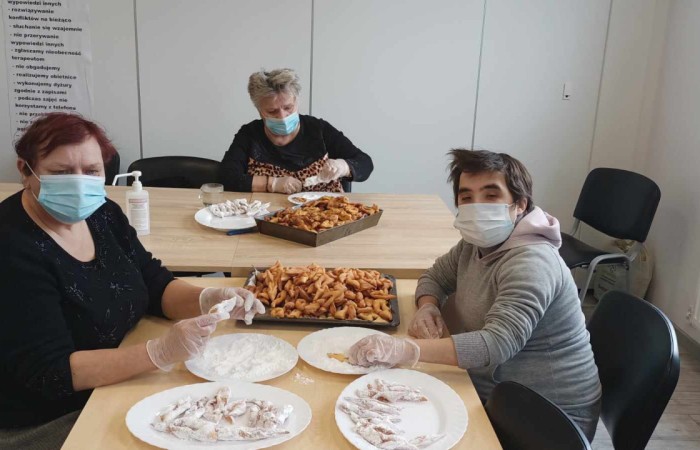 Trzy osoby w maseczkach i rękawiczkach przygotowują słodkie wypieki przy dużym stole. Na pierwszym planie kobieta w czarnej bluzce i mężczyzna w szarej bluzie obtaczają ciastka w cukrze pudrze, obok nich leżą talerze z gotowymi wypiekami. W tle starsza kobieta przygotowuje kolejne porcje na tacy.