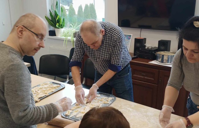 Troje ludzi pracuje wspólnie przy stole nad przygotowaniem faworków. Mężczyzna w koszuli w kratę i kobieta z długimi ciemnymi włosami układają kawałki ciasta na blasze. Obok nich stoi mężczyzna w okularach. 