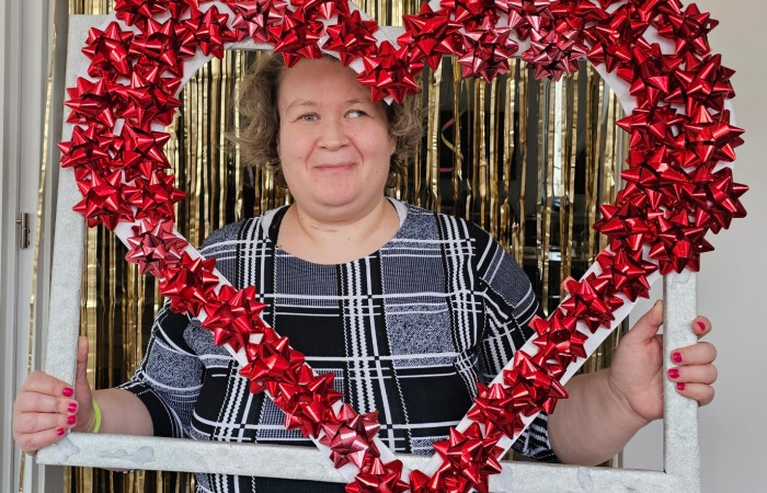 Kobieta w czarno-białej sukience z krótkimi, kręconymi włosami uśmiecha się, trzymając dekoracyjną ramkę w kształcie serca ozdobioną czerwonymi kokardkami. W tle złote, połyskujące zasłony.