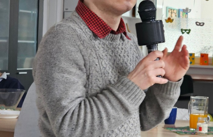 Ten sam mężczyzna w czerwonej koszuli, tym razem w szarym swetrze, stoi w sali, trzymając mikrofon i przemawiając. W tle stoliki, szklanki z napojami i dekoracje na ścianach.