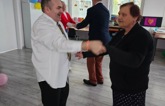 Mężczyzna w białej koszuli i kobieta w czarnej sukience z białym wzorem tańczą razem w udekorowanej sali. W tle inni uczestnicy tańca oraz kolorowe balony przyczepione do ściany.