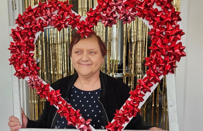 Ta sama kobieta w czarnej sukience pozuje z dekoracyjną ramką w kształcie serca, uśmiechając się do aparatu. W tle złote zasłony