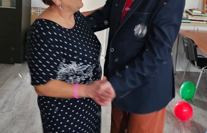Starszy mężczyzna w granatowej marynarce i czerwoną krawatą tańczy z kobietą w czarnej sukience. Oboje trzymają się za ręce i uśmiechają. W tle kolorowe balony i dekoracje karnawałowe.