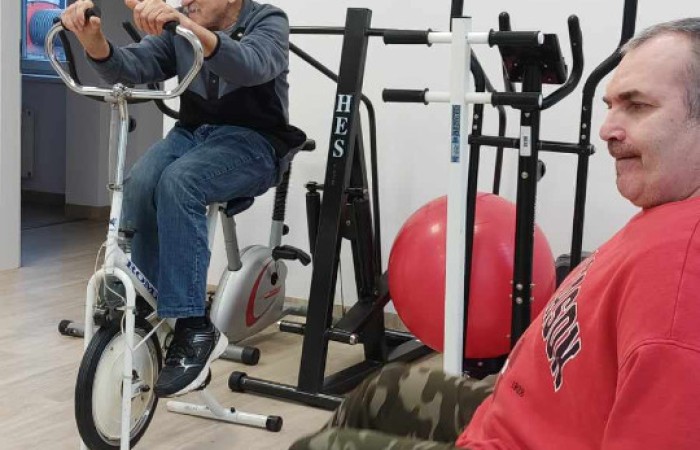 Dwóch mężczyzn korzysta ze sprzętu do ćwiczeń w sali rehabilitacyjnej. Jeden starszy mężczyzna w ciemnej kamizelce i jeansach pedałuje na rowerku stacjonarnym, a drugi, ubrany w czerwoną bluzę i spodnie moro, siedzi na krześle i korzysta z mini rowerka do ćwiczeń nóg. W tle sprzęty sportowe i dekoracje ścienne. 
