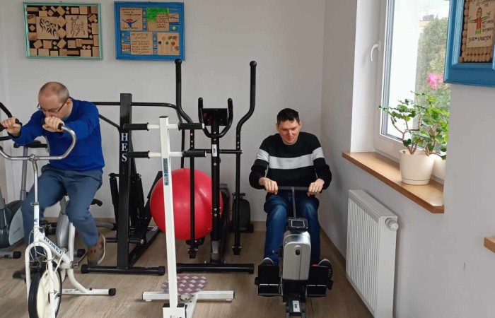 Dwóch mężczyzn ćwiczy na sprzęcie treningowym w jasnym pomieszczeniu. Jeden pedałuje na rowerku stacjonarnym, a drugi siedzi przy wioślarzu i wykonuje ruchy wiosłowania. W tle znajdują się przyrządy sportowe, czerwone piłki rehabilitacyjne i okna z widokiem na zewnątrz.  