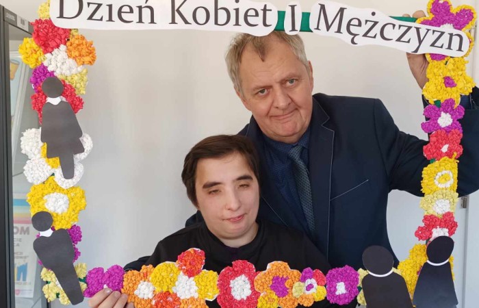 Mężczyzna w ciemnym garniturze i młoda osoba w czarnym ubraniu pozują do zdjęcia, trzymając dekoracyjną ramkę z napisem „Dzień Kobiet i Mężczyzn”. W tle przeszklona witryna z nagrodami i pucharami.