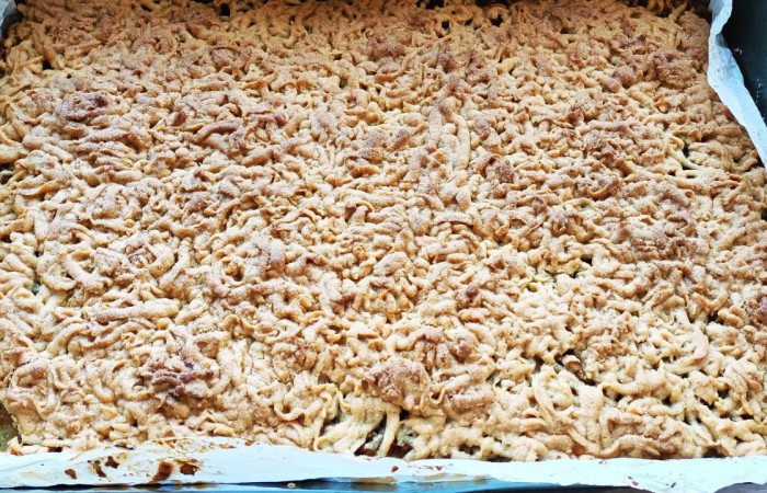 Blacha z upieczonym ciastem o złocistym kolorze i kruszonkowej strukturze. Ciasto znajduje się na papierze do pieczenia, lekko wystaje poza krawędzie formy. 