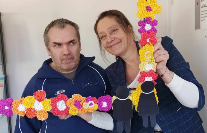 Mężczyzna w granatowej bluzie i kobieta w ciemnym żakiecie trzymają dekoracyjną ramkę „Dzień Kobiet i Mężczyzn”. Kobieta uśmiecha się szeroko, mężczyzna patrzy poważnie.