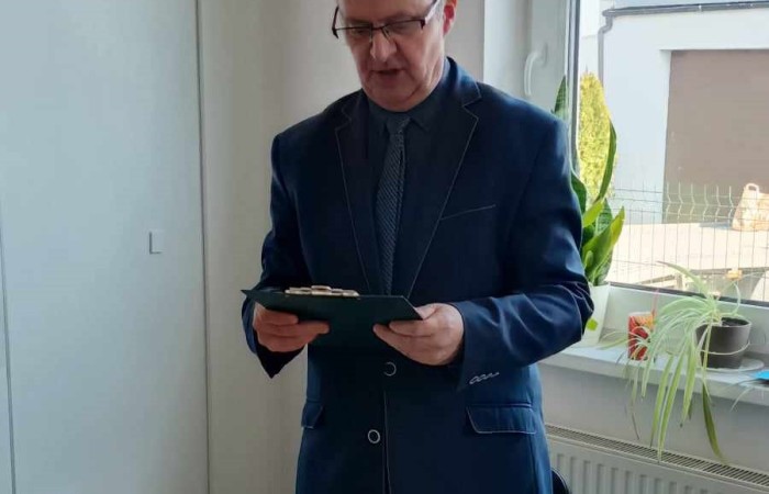 Mężczyzna w eleganckim ciemnym garniturze i krawacie stoi przy stole, trzymając kartkę i czytając na głos. Na stole leży czarna filiżanka i talerz z resztkami ciasta. W tle okno z roślinami. 