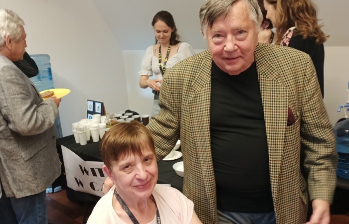 Zdjęcie starszej pani siedzącej na wózku ma na sobie różową koszulkę, przypinkę z logiem i smycz z plakietką FFnC, na kolanach trzyma torebkę. Obok niej stoi aktor ma na sobie żółtą marynarkę w kratę, czarną koszulkę i ciemne spodnie. Są uśmiechnięci. W tle widać bufet.
