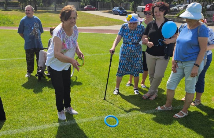 Kolejna osoba rzucająca plastikową obręcz do kolorowych palików na trawie, otoczona przez grupę seniorów.
