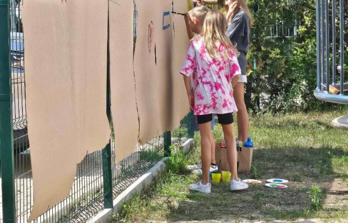Trzy dziewczynki stoją przy ogrodzeniu i malują na dużych arkuszach kartonu, które są przypięte do płotu. Obok leżą farby i pędzle