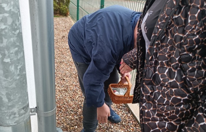 Osoba w granatowej kurtce schyla się, trzymając mały koszyk z przezroczystą torebką wypełnioną przedmiotami. Obok stoi kobieta w czarnej kurtce z jasnymi włosami. Teren pokryty drobnym żwirem, w tle widoczny zielony płot i drzewa