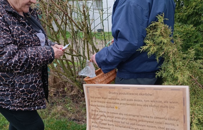 Kobieta w czarnej kurtce i mężczyzna w granatowej kurtce oraz szarej czapce stoją przy krzewie. Mężczyzna trzyma koszyk, a kobieta coś zapisuje. Na pierwszym planie widoczna jest kartka z instrukcją, lekko wygięta.