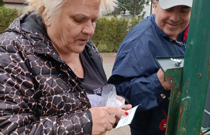 Kobieta w czarnej kurtce trzyma kartkę i foliową torebkę z kolorowymi przedmiotami. Obok niej mężczyzna w granatowej kurtce otwiera metalową skrzynkę na zielonym ogrodzeniu. W tle drzewa i krzewy kobieta i mężczyzna stoją w pobliżu krzewów, trzymając koszyk wypełniony przedmiotami zapakowanymi w foliowe torebki.