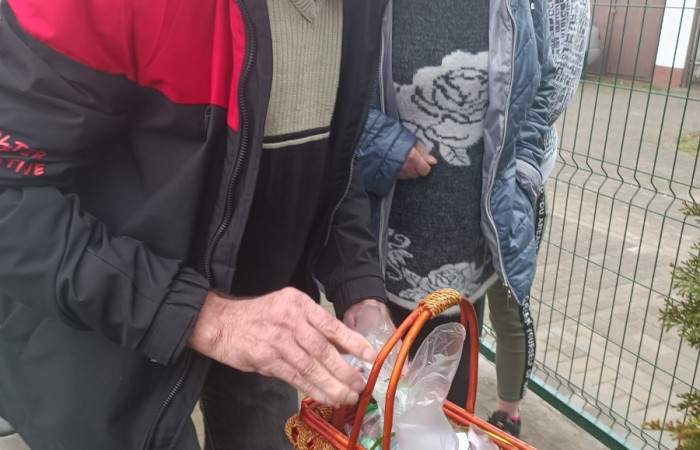 Starszy mężczyzna w czapce i czarno-czerwonej kurtce trzyma wiklinowy koszyk wypełniony foliowymi torebkami. Za nim stoją dwie osoby, jedna z nich ma szarą kurtkę z wzorem róży